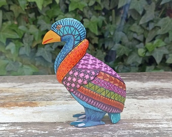 May include: A colorful wooden bird sculpture with intricate geometric patterns in shades of blue, orange, pink, green, and brown. The bird is standing on a wooden surface.