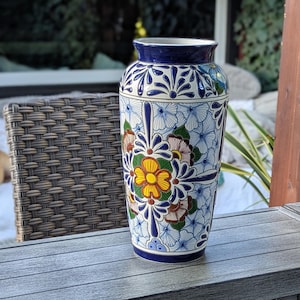 May include: A tall, ceramic vase with a vibrant floral design. The vase features a blue rim and base, with a white body adorned with blue, yellow, and green floral patterns. The vase is displayed on a gray surface.