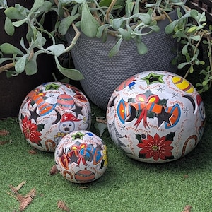 May include: Three spherical ceramic garden ornaments of varying sizes, each featuring a white base with hand-painted Christmas motifs including red poinsettias, orange bells, snowmen, and black stars. Each sphere has circular cutouts.