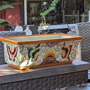 May include: A rectangular ceramic planter featuring a cream-colored base with a hand-painted design of colorful butterflies and floral motifs. The rim and base are finished in a solid orange glaze, providing a vibrant contrast to the detailed pattern.