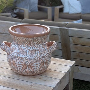 May include: A terracotta-colored ceramic vase with a wide opening and two handles. The vase features a white, intricate floral pattern. The vase is sitting on a wooden table.