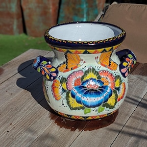 May include: A hand-painted ceramic planter with a white base, decorated with colourful floral and butterfly designs. The planter has a blue rim and handles, with a vibrant blue, yellow, and orange floral pattern. It is on a wooden surface.