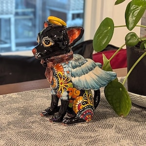 May include: A ceramic figurine of a black dog with a yellow halo, angel wings, and a colorful patterned coat. The dog has a red nose and ears. The figurine is sitting on a patterned surface.