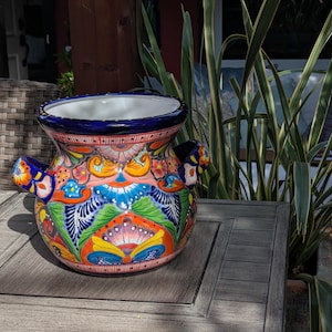 May include: A vibrantly painted ceramic vase with a wide, open top and two small handles. The vase features a colorful floral design with blue, orange, green, and yellow hues. The rim is a deep blue, and the interior is white.