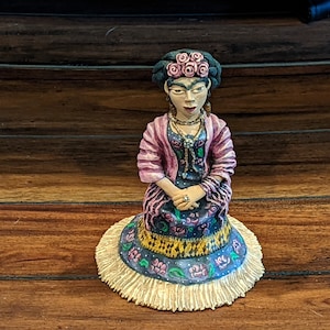 May include: A seated figurine of a woman with dark hair and pink rose hair decoration. She wears a pink and purple striped jacket over a floral dress. The figurine is on a round base with a fringed edge, hands clasped.