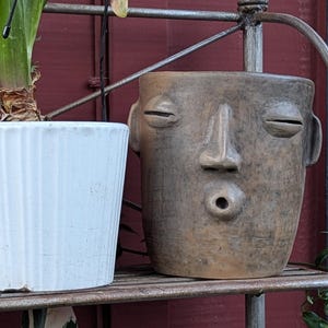 May include: Two decorative planters on a metal shelf. A white, ribbed ceramic planter sits next to a brown, face-shaped planter. The face planter has closed eyes and a round mouth. The shelf is against a dark red and black background.