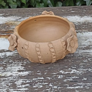 May include: A terracotta bowl with a decorative floral design. The bowl has a rounded shape and a smooth, unglazed finish.
