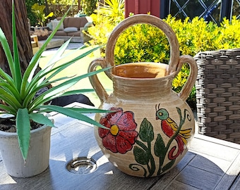 May include: A hand-painted terracotta pot with a floral design and a bird. The pot has a handle and two side handles. The pot is sitting on a wooden table.