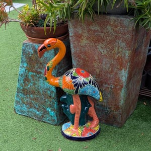 May include: A colorful ceramic flamingo statue with an orange neck and head, and a body adorned with blue, orange, and yellow floral patterns. The flamingo stands on a green base with a similar floral design. The statue is placed on green artificial turf.