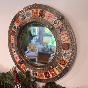 May include: Round decorative mirror with a silver metal frame and colorful square tile accents. The mirror reflects a room with furniture and natural light. The tiles feature various patterns in red, orange, blue, and yellow.