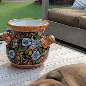 May include: A hand-painted ceramic vase with a black background and colorful floral patterns. The vase has two handles and a wide opening. The rim and handles are accented with orange and yellow. The vase is sitting on a wooden surface.
