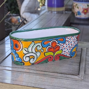 May include: A rectangular ceramic planter with a vibrant, hand-painted design. The planter features a white interior, a green rim, and a colorful exterior with floral and geometric patterns in orange, blue, yellow, and red. A decorative piece for plants.