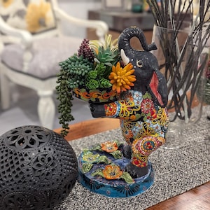 May include: A decorative elephant figurine with a colorful floral design, holding a basket of succulents and flowers. The elephant is black with red and white accents. A black, patterned sphere is in the foreground.