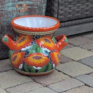 May include: A hand-painted ceramic vase with a wide opening and two handles. The vase features a vibrant floral design with orange and yellow flowers on a blue and white background. The rim and handles are orange.