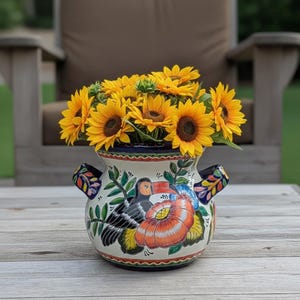 May include: A ceramic vase with a colorful floral and bird design, filled with bright yellow sunflowers. The vase has two handles and is sitting on a wooden table. The background is blurred, showing a chair and greenery.