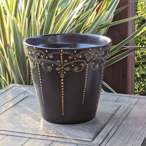 May include: A black ceramic planter with gold painted details and a decorative design. The planter is sitting on a wooden surface.