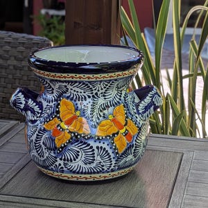 May include: A blue and white ceramic planter with a floral design and two orange butterflies. The planter has a wide mouth and two handles.
