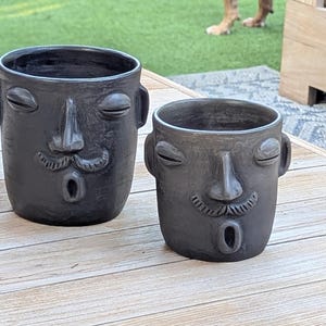 May include: Two dark gray ceramic planters with human face designs. Each planter has closed eyes, a nose, a mustache, and an open mouth. The planters are on a light wood surface.