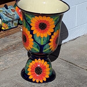 May include: A tall, hand-painted ceramic planter with a flared top and pedestal base. The planter is adorned with vibrant orange and yellow sunflowers against a dark blue background. The interior is a light cream color, and the overall design is a bold, decorative piece.