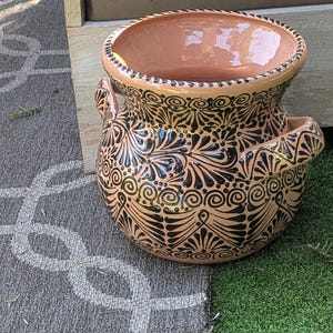 Op de afbeelding: Een terracotta plantenpot met een zwart en bruin bloemenmotief. De pot heeft twee handvatten aan de zijkant en een brede opening aan de bovenkant.