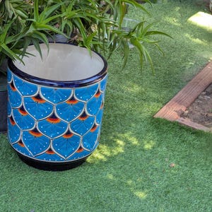May include: A cylindrical ceramic planter with a blue, orange, and white scale pattern. The planter has a black base and a dark blue rim. The interior is white. The planter is outdoors, surrounded by green grass and plants.