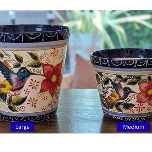 May include: Two ceramic flower pots with a hand-painted design. The pots feature a cream-colored base with a hummingbird and floral pattern in red, yellow, blue, and green. The rims are dark blue. The larger pot is labeled "Large" and the smaller pot is labeled "Medium".
