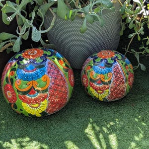 May include: Two spherical garden ornaments featuring a vibrant, hand-painted Talavera-style pattern. The design includes intricate floral motifs and geometric shapes in bright shades of red, orange, yellow, blue, green, and black on a glossy surface.