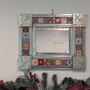 May include: Square silver metal framed mirror with decorative tile accents. The frame has embossed floral designs at the corners and along the sides. The tiles feature colorful floral patterns.