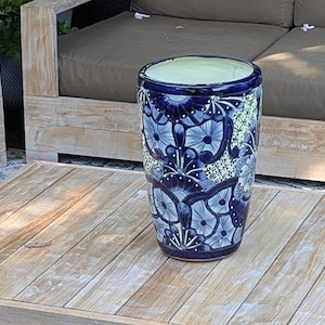 May include: A cylindrical ceramic vase featuring a deep blue and white floral pattern with intricate hand-painted details. The vase has a smooth, glossy finish, a wide circular opening, and a tapered base, showcasing a complex botanical design.