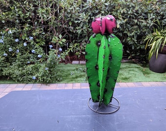 May include: A metal sculpture of a cactus with a pink flower on top. The cactus is green and has a textured surface. The sculpture is on a black metal stand.