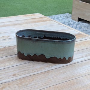 Könnte beinhalten: Ein ovaler Keramik-Pflanztopf mit zweifarbigem Glanz. Der obere Rand ist dunkelbraun, geht in ein türkisgrün über und hat dann einen dunkelbraunen Sockel. Der Pflanztopf steht auf einem hellen Holztisch.