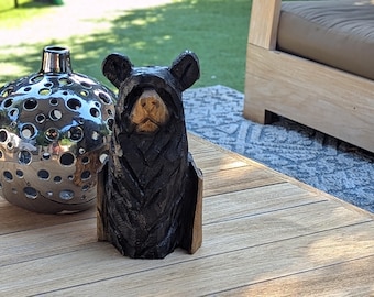May include: A hand-carved wooden bear sculpture with a black body and a natural wood-colored face. The bear is sitting upright. A silver decorative vase with circular cutouts is to the left of the bear.