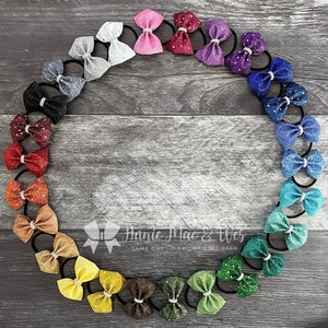 May include: A set of 20 glitter hair bows with rhinestones in a variety of colors, arranged in a circle on a woodgrain background. The bows are all tied with black elastic hair ties. The text "Auntie Mac & Wes GAME DAY SIDELINE ALL STAR" is visible at the bottom of the image.