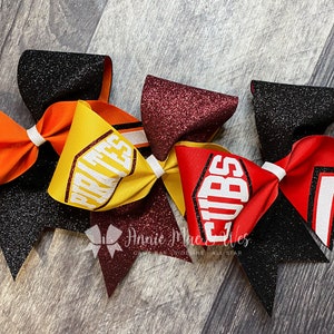 May include: A collection of cheerleading bows in various colors, including orange, yellow, red, and black. Each bow features glitter accents and team names like "PIRATES" and "CUBS" in bold white lettering. The bows are tied with white ribbon.
