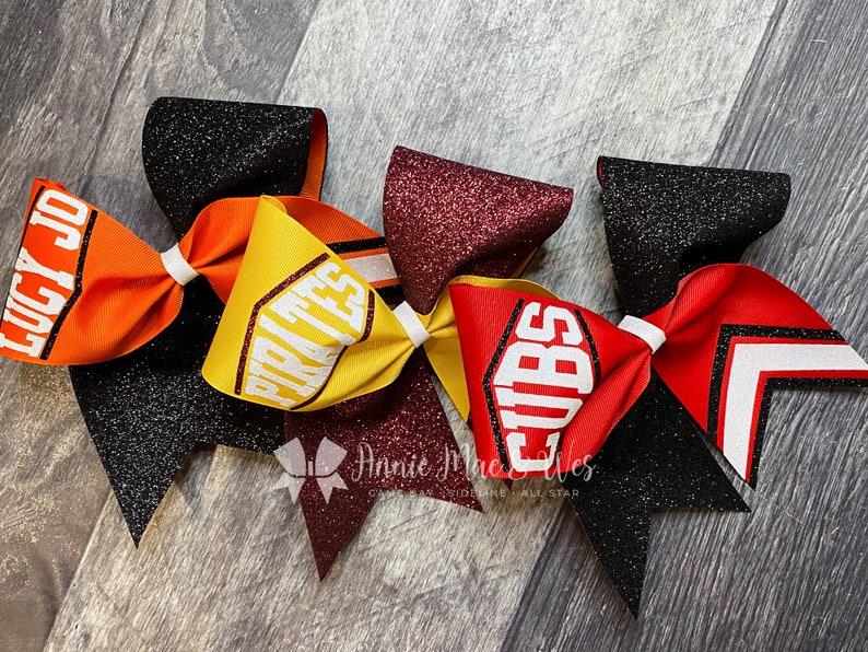 May include: A collection of cheerleading bows in various colors, including orange, yellow, red, and black. Each bow features glitter accents and team names like "PIRATES" and "CUBS" in bold white lettering. The bows are tied with white ribbon.
