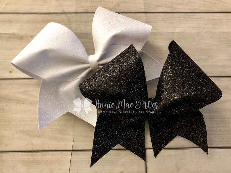 May include: Two glittery hair bows. One is white, the other is black. The bows are layered on top of each other. The text "Annie Mac & Wes" is visible on the bottom right of the image.