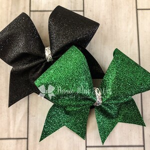 May include: Two glittery cheerleading bows. One is black, the other is green. Each bow has a silver rhinestone center. The bows are on a light wood grain background. The bows are likely for a school or team.