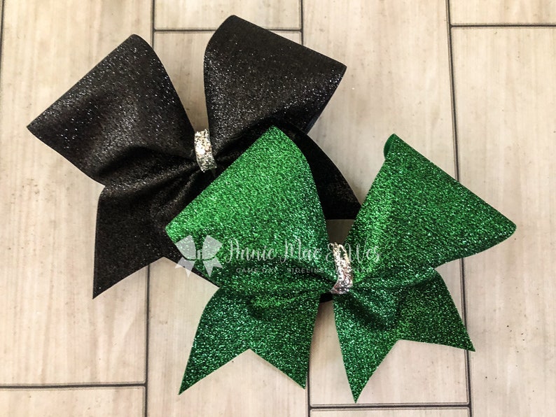 May include: Two glittery cheerleading bows. One is black, the other is green. Each bow has a silver rhinestone center. The bows are on a light wood grain background. The bows are likely for a school or team.