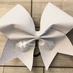 May include: A large, white glitter cheerleading bow with a center knot and elastic band. The bow has a sparkly texture and is designed for game day or all-star events. The Annie Mac & Wes logo is visible.