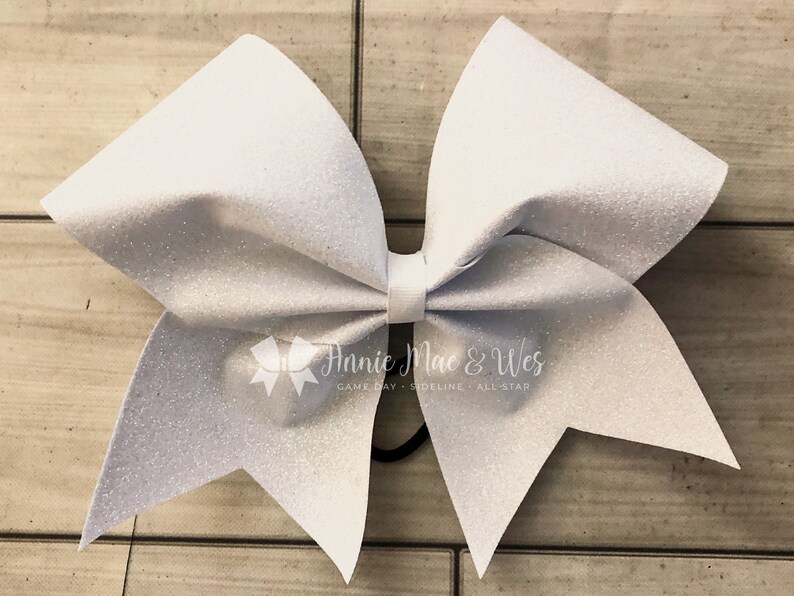 May include: A large, white glitter cheerleading bow with a center knot and elastic band. The bow has a sparkly texture and is designed for game day or all-star events. The Annie Mac & Wes logo is visible.