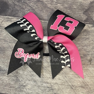 May include: Black and pink cheer bow with white stitching and the number "13" in white on a pink background. The bow also has the name "Buford" in white on a black background.