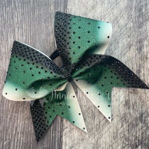 May include: A large cheerleading bow with a gradient design, transitioning from white to green and black. The bow is embellished with black rhinestones and features a black center knot. The text "GAME DAY" is visible.