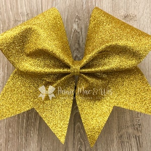 May include: A large, gold glitter cheerleading bow. The bow is made of a shiny, textured material and has a classic bow shape. The bow has a small white logo that says "Annie Mac & Wes".