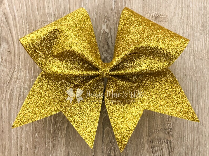 May include: A large, gold glitter cheerleading bow. The bow is made of a shiny, textured material and has a classic bow shape. The bow has a small white logo that says "Annie Mac & Wes".