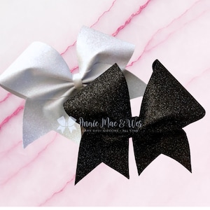 May include: Two glittery cheerleading bows. One is silver, the other is black. The bows are on a pink marble background. The text "Annie Mac & Wes" is printed on the black bow.