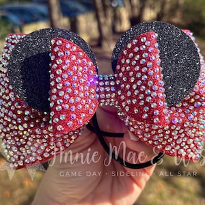 May include: A red and black bow with sparkling rhinestones. The bow features a black glitter circle and red bow sections, all adorned with clear rhinestones. The bow is held together with a black elastic band. The text "Annie Mae & Wes" is visible.