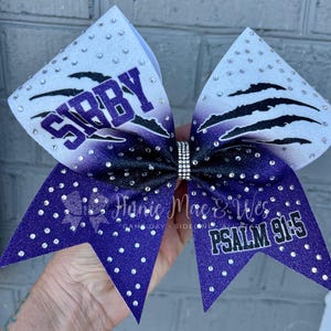 May include: Purple and white cheer bow with silver glitter and rhinestones. The bow has the name "SIBBY" on one side and "PSALM 91:5" on the other side.