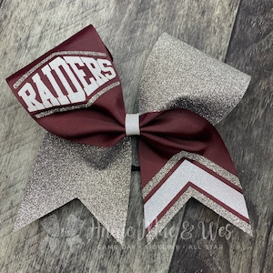 May include: A maroon and silver cheer bow with the word "RAIDERS" in white letters on the maroon section. The bow has a silver glitter finish.
