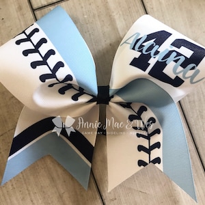 May include: A white, blue, and navy blue cheer bow with a baseball stitch design and the name "Aunna" and the number "42" printed on it.