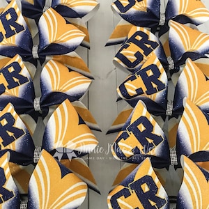 May include: A collection of cheerleading bows. Each bow features a navy blue and gold color scheme with the letters "CR" in gold. The bows have a glittery finish and a rhinestone center.